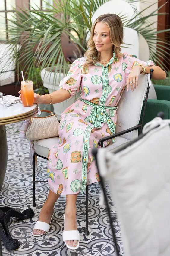 A woman wearing a pink dress is seated on a chair, enjoying a vibrant orange drink.